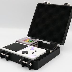 May include: Black, hard-shell carrying case, open to reveal a white and purple handheld gaming device and a stack of game cartridges. The case has a black interior and silver latches. A retro gaming theme.