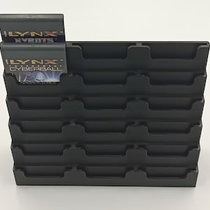 May include: A black plastic game cartridge holder with multiple slots. Two game cartridges are inserted, displaying the text "LYNX" and "X-bots" on one, and "CYBERBALL" on the other. The holder is designed to store multiple game cartridges.