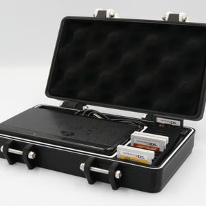 May include: Black hard-shell carrying case, open to reveal a black handheld gaming device, charging cable, and game cartridges. The case has a foam interior and silver latches. The case is designed to protect the gaming device and accessories.