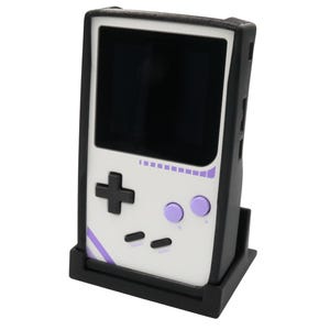 May include: A handheld video game console with a white faceplate, a black screen, a black directional pad, two light purple action buttons labeled A and B, and two black buttons labeled SELECT and START, resting in a black plastic stand.