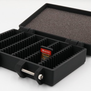 May include: Black, open, hinged case with multiple vertical slots for game cartridges. The case has a foam-lined lid and metal bars on the front. Two red and black game cartridges are visible in the slots. The case is designed to hold video game cartridges.