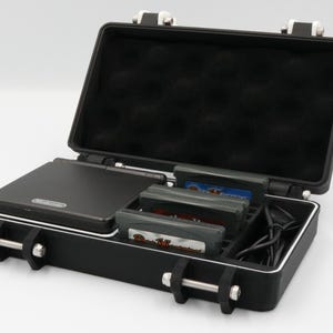 May include: Black case with a white trim, open to reveal a black Nintendo Game Boy Advance SP, game cartridges, and a charging cable. The case has a foam interior and metal latches.