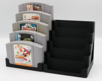 May include: A black plastic video game cartridge holder with five gray game cartridges. The cartridges are stacked and display colorful game labels. The holder has a separate section with four slots for additional game storage.
