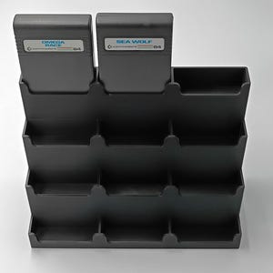 May include: A black, multi-tiered organizer designed to hold video game cartridges. Two cartridges labeled "Omega Race" and "Sea Wolf" are visible. The organizer is made of a matte black material and has multiple slots for storage.