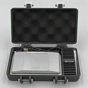 May include: An open, black, hard-shell case with a silver Nintendo DS Lite inside. The case has a foam interior and metal latches. A game cartridge and stylus are also visible.