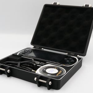 May include: Black hard case with a black interior, holding a black handheld gaming console, a disc, and a charging cable. The case has silver latches and hinges, and the interior is lined with black foam.