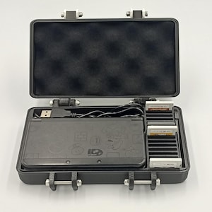 May include: Black Nintendo 3DS console with a Mario-themed design, inside a black carrying case. The case is open, revealing the console, game cartridges, and a USB cable. The case has silver latches and hinges.