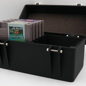 May include: Black storage case with a hinged lid, open to reveal a compartment with a stack of video game cartridges. The case has metal latches and a handle. The cartridges have colorful labels.