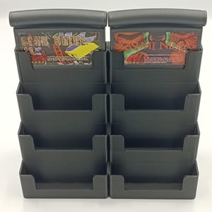 May include: A black plastic video game cartridge holder with multiple slots. Two game cartridges are visible, one labeled "Club Drive" and the other "Kasumi Ninja". The holder is designed to organize and display video game cartridges.