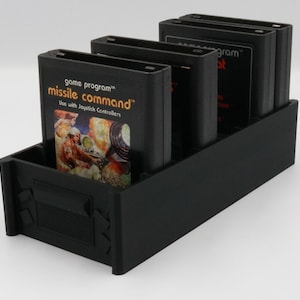 May include: Black plastic game cartridge holder with several black game cartridges. The front cartridge displays "Missile Command" with a colorful graphic. The holder is rectangular with a slot for the cartridges.