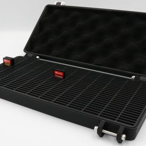 May include: Black plastic case with an open lid. The interior has a black foam lining and a grid-like tray. Two small red and black labels are visible on the tray. The case has silver hinges and latches.