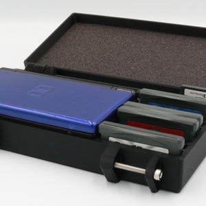 May include: Black carrying case for a Nintendo DS, open to reveal a blue DS console and several game cartridges. The case has a foam interior and secure latches for travel. A DS game is visible.