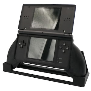 May include: A black handheld gaming console with dual screens, a directional pad, and action buttons labeled X, Y, A, and B. The device is mounted in a black plastic ergonomic grip stand that features side handles for improved comfort during gameplay.