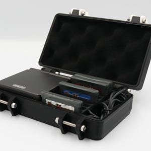 May include: Black carrying case with a hinged lid, holding a gray handheld game console, game cartridges, and a charging cable. The interior is lined with black foam padding. The case has silver latches and hinges.