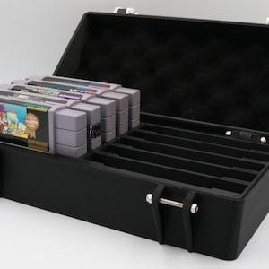 May include: Black storage case with silver latches, open to reveal a compartment for video game cartridges. The case contains several gray game cartridges, with colorful labels. The interior of the lid is lined with black foam.
