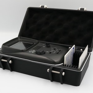 May include: Black hard case with a built-in electronic device and a compartment for cards. The case is open, revealing a screen, buttons, and a control knob. The interior is lined with black foam.