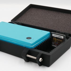 May include: Black protective case with a teal handheld gaming console inside. The case is open, revealing a game cartridge labeled "Rooms" and a foam-lined lid. The case has metal latches.