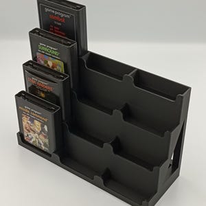May include: A black plastic video game cartridge organizer with multiple slots. The organizer holds several game cartridges, including "Combat," "Surround," "Star Raiders," and "Missile Command." The design features tiered levels for easy access and display.