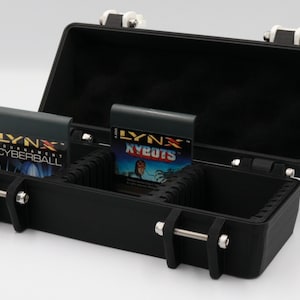 May include: Black case with silver latches and hinges, open to reveal two Lynx game cartridges. The cartridges are labeled "LYNX" and feature game titles "CYBERBALL" and "NYEUTS". The case is designed to hold and protect the game cartridges.