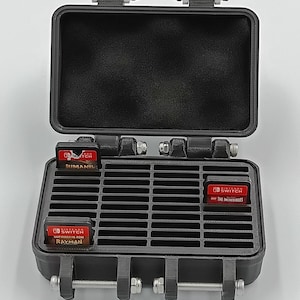 May include: Black, open, rectangular case with a foam interior and a grid of slots for game cartridges. The case is secured with latches and hinges. Three red Nintendo Switch game cartridges are visible, labeled "Mimami", "Rayman", and "The Incredibles".