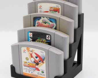 May include: A black plastic game cartridge holder with five gray Nintendo 64 game cartridges. The cartridges have colorful labels with game titles and graphics. The top cartridge reads "NFL Quarterback Club."