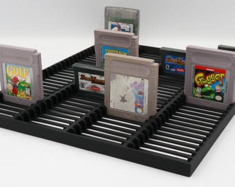 May include: A black plastic game cartridge organizer holding several vintage video game cartridges. The cartridges are in various colors and feature game labels. The organizer has a slotted design for easy cartridge access and display.