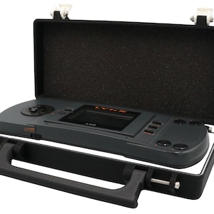 May include: A dark gray handheld video game console with the word LYNX printed in orange above the screen. The console is housed inside a black rectangular carrying case with a black handle and foam lining on the interior of the lid.