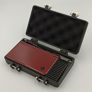 May include: A burgundy handheld gaming console inside a black case with a foam interior. The case is open, revealing the console and a storage compartment. The case has silver latches and hinges.