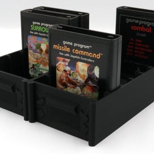 May include: A black plastic organizer holding several Atari game cartridges. The cartridges are labeled "Surround", "Missile Command", and "Combat". The organizer has multiple compartments and is designed to hold the game cartridges upright.