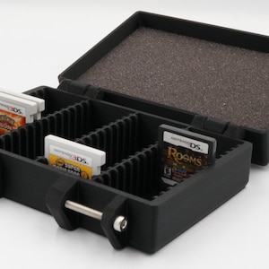 May include: Black plastic game cartridge carrying case, open to reveal game cartridges. The case has a hinged lid with a foam interior. The case is designed to hold multiple game cartridges, with dividers to keep them organized.