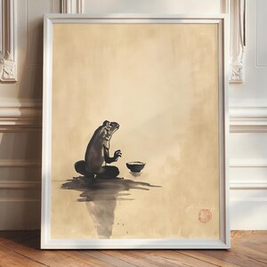 May include: A framed art print featuring a black and white ink wash painting of a frog in a meditative pose. The frog sits cross-legged with a small bowl nearby, set against a beige background. A red circular stamp is in the lower right corner.