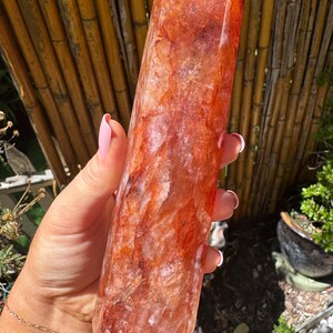 May include: A large, polished crystal tower in shades of reddish-orange. The crystal has a faceted top and a smooth, cylindrical body. The stone is held in a hand, with a bamboo background and greenery visible.