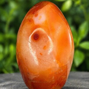 May include: A polished carnelian gemstone, oval-shaped, with a vibrant orange hue. The stone features a lighter, swirling pattern with a central, eye-like inclusion. The surface is smooth and reflective, showcasing natural variations in color and texture.