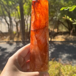 May include: A tall, orange-red crystal point with a faceted top. The crystal has a translucent quality, with variations in colour and texture. The crystal is held in a hand against a blurred green background of foliage.