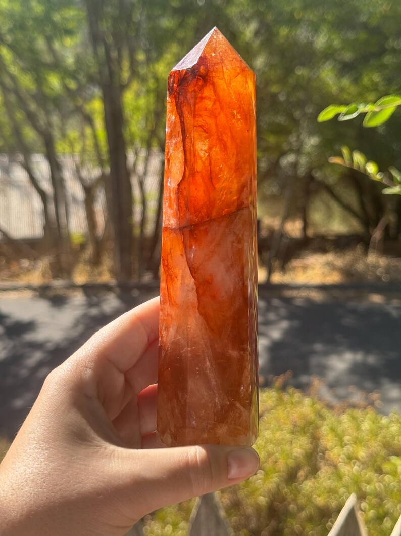 May include: A tall, orange-red crystal point with a faceted top. The crystal has a translucent quality, with variations in colour and texture. The crystal is held in a hand against a blurred green background of foliage.