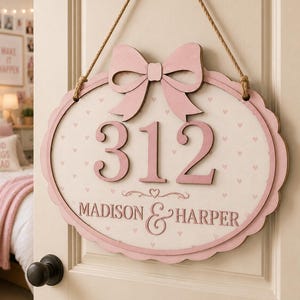 May include: An oval wooden door sign featuring a light pink bow at the top and the number 312 in large pink digits. The background is cream with a small pink heart pattern. Below the number, the text reads MADISON & HARPER in pink lettering.