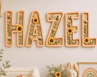 Puede incluir: Un conjunto de cinco letras de madera para pared que forman el nombre HAZEL, con un borde color melocotón claro y un interior de madera natural. Cada letra está decorada con girasoles tridimensionales, flores blancas, hojas verdes y abejas.