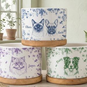 Puede incluir: Tres macetas de cerámica para mascotas con bases de bambú. Cada maceta presenta un retrato de mascota diferente: un gato siamés y un bulldog francés, un gato esponjoso y un Border Collie. Patrones florales en azul, morado y verde rodean los retratos.