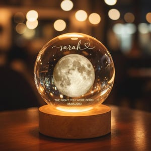 Peut inclure: Une sphère de verre transparente avec une image détaillée de la lune à l'intérieur, posée sur une base en bois qui émet une lueur chaude. La sphère est inscrite du nom "sarah" et de la phrase "THE NIGHT YOU WERE BORN 08.06.2010". L'arrière-plan est flou avec des lumières bokeh.