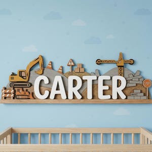 Puede incluir: Letrero de nombre de madera con el nombre "CARTER" en letras blancas, con temática de construcción. Incluye una excavadora amarilla, una excavadora, una grúa y otros elementos de construcción. El letrero está montado en una estantería de madera sobre un fondo azul.