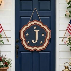 Könnte beinhalten: Eine marineblaue Eingangstür mit einem dekorativen Holzschild mit gewelltem Rand, einem zentralen Buchstaben J und fünf Sternen. Die Tür wird von zwei amerikanischen Flaggen, Topfblumen, einer Wandlaterne und einer Fußmatte flankiert.