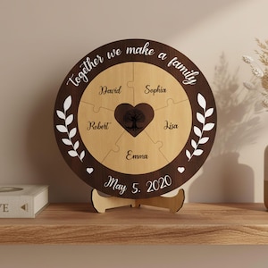Può includere: Un puzzle di famiglia in legno con la scritta "Together we make a family". Il puzzle ha sei pezzi, con nomi e un centro a forma di cuore con un disegno di albero. È inclusa anche la data "May 5. 2020".