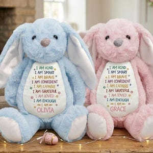 Può includere: Due coniglietti di peluche, uno azzurro chiaro e uno rosa, sono affiancati. Ognuno ha lunghe orecchie e una pancia bianca con testo ricamato: "I AM KIND, I AM SMART, I AM BRAVE, I AM CONFIDENT, I AM CAPABLE, I AM GRATEFUL, I AM LOVED, I AM ENOUGH." Il coniglietto blu ha il nome "OLIVIA", e quello rosa ha il nome "GRACE".