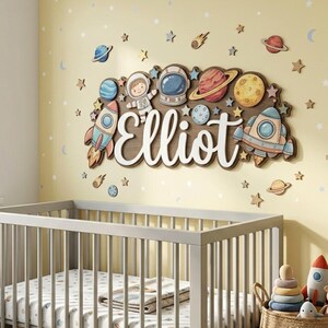 Könnte beinhalten: Wanddekoration für das Kinderzimmer mit dem Namen "Elliot" in Schreibschrift, umgeben von bunten Planeten, Raketen, Astronauten und Sternen. Die Dekoration befindet sich an einer gelben Wand mit Stern- und Mondakzenten, über einem grauen Babybett.