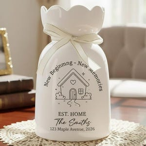May include: A white ceramic vase with a scalloped top and a cream-colored ribbon tied around the neck. The vase features a house graphic and the text "New Beginning - New Memories" and "EST. HOME The Smiths 123 Maple Avenue, 2026".