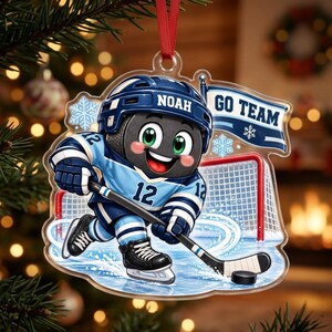 May include: A clear acrylic Christmas ornament featuring a cartoon hockey puck character wearing a blue and white jersey with the number 12 and a helmet labeled NOAH. The character holds a hockey stick near a net and a flag reading GO TEAM.