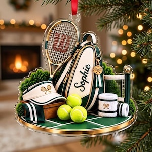 May include: A tennis-themed Christmas ornament featuring a racket, a tennis bag personalized with the name Sophie, a visor, three neon yellow tennis balls, and two white wristbands with green stripes, all arranged on a green court base.
