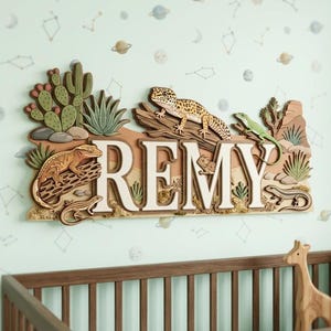 Puede incluir: Un letrero de pared de madera multicapa con el nombre REMY en letras blancas grandes. El diseño incluye varios lagartos, como un gecko leopardo y un gecko crestado, junto a plantas del desierto como cactus sobre un fondo color canela.