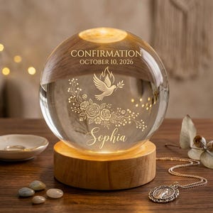 Può includere: Una sfera di vetro trasparente con il testo "CONFIRMATION OCTOBER 10, 2026" e il nome "Sophia" inciso. La sfera presenta un design a colomba e floreale e poggia su una base di legno. Un piattino e una collana sono in primo piano.