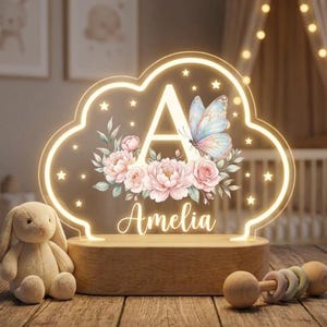 May include: A cloud-shaped LED night light with a wooden base, featuring a large letter A, a colorful butterfly, and pink floral illustrations. The name Amelia is printed in white script below the flowers, surrounded by small glowing stars.